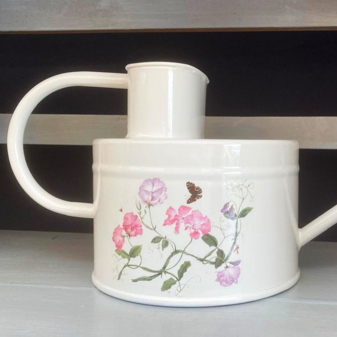 A white watering can with off white paint and a floral Sweet Pea design, including pink flowers and butterflies, painted on the side.