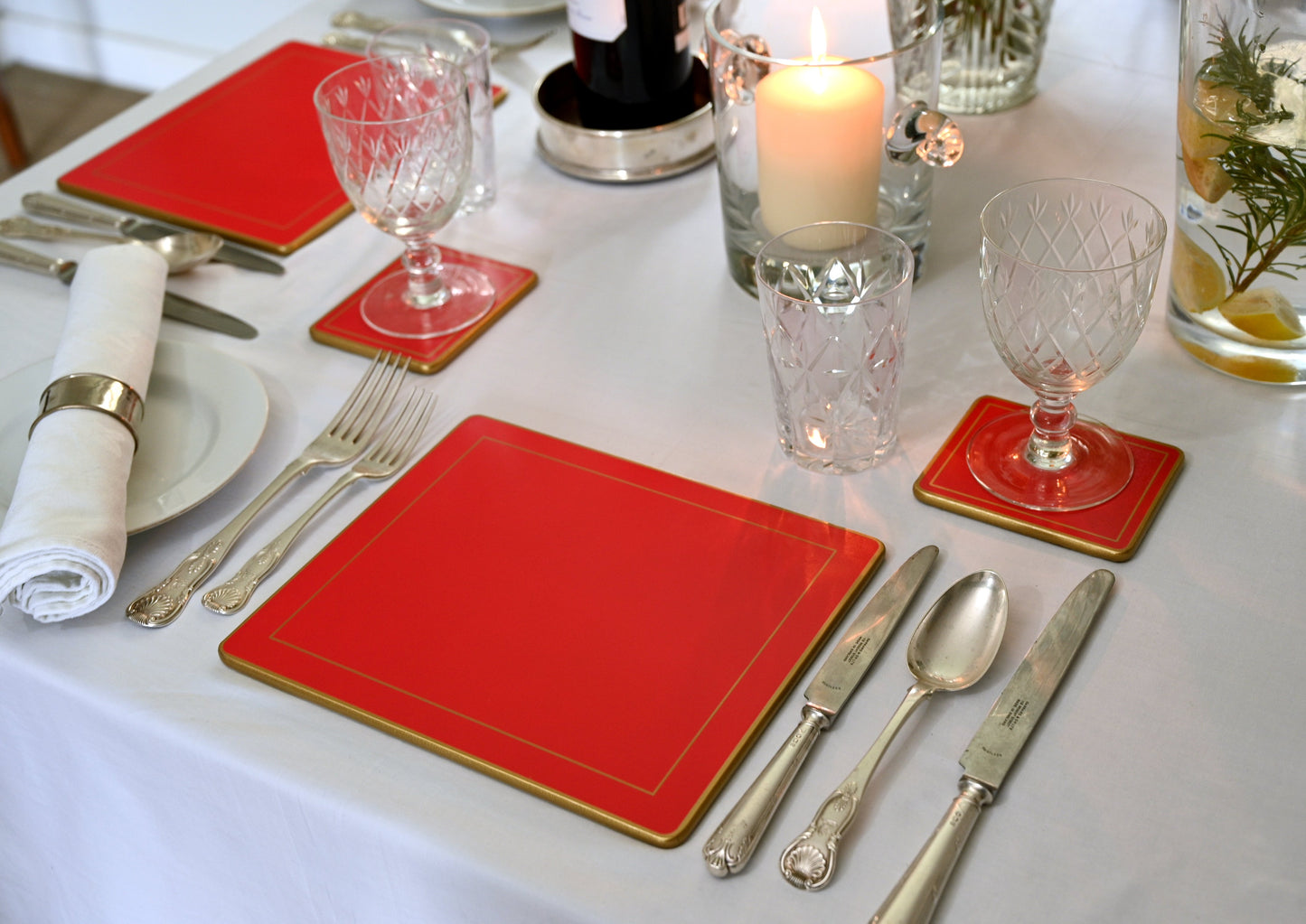 Dining table setting with red placemats, silverware, and glasses on a white tablecloth.