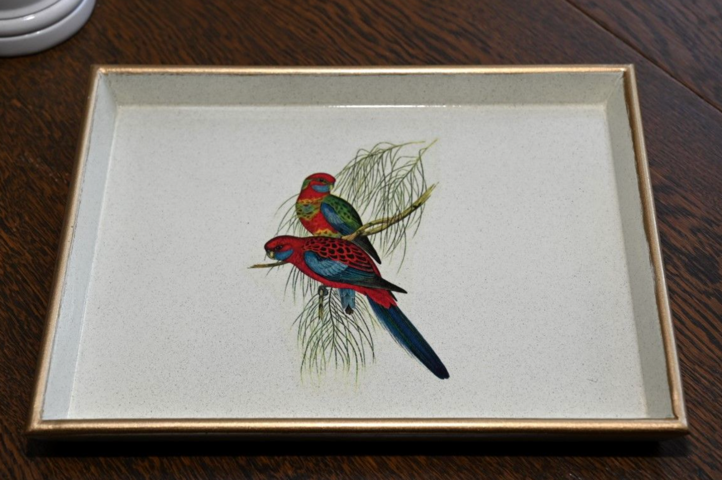 A wooden lacquered tray with a gilded gold rim, featuring an illustration of a pair of parrots perched on a branch, on a wooden table background